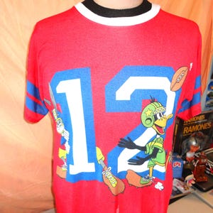 May include: Red vintage football jersey with the number 12 in blue and white. The jersey features a cartoon duck character in a football uniform. The sleeves have blue and red stripes. The collar is white and black.