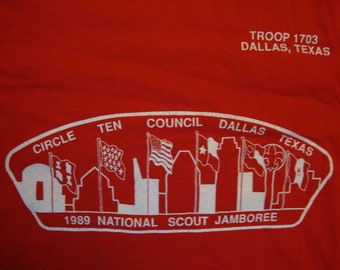 May include: White text on a red shirt reads "TROOP 1703 DALLAS, TEXAS" and "CIRCLE TEN COUNCIL DALLAS TEXAS 1989 NATIONAL SCOUT JAMBOREE". The text is in a curved banner shape with a city skyline and flags.