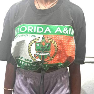 May include: A charcoal gray short-sleeve t-shirt featuring a large graphic print that reads FLORIDA A&M UNIVERSITY HOMECOMING 1996 RATTLERS. The design includes a central crest with a torch, a book, and a snake, surrounded by green and orange accents.