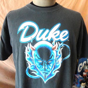 Pode incluir: Uma t-shirt cinzento-carvão de manga curta com a palavra Duke em letras brancas e um contorno azul-claro. Abaixo do texto, há um gráfico de um rosto de diabo azul rodeado por chamas azuis, com pequenos chifres e uma cauda visível.
