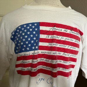 Op de afbeelding: Een wit T-shirt met een vintage Amerikaanse vlag-afbeelding en de tekst "America the Beautiful" met songteksten. De tekst luidt: "Oh beautiful for spacious skies, for amber waves of grain, for purple mountain majesties above the fruited plain. America, America, God shed His grace on thee, and crown thy good with brotherhood, from sea to shining sea." Onderaan het shirt staat "Cape Cod".