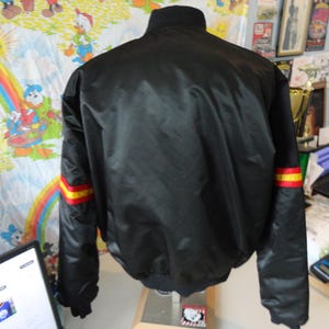 May include: Black bomber jacket with a ribbed collar and cuffs. The sleeves have red and yellow stripes near the cuffs. The jacket is made of a shiny material. The back of the jacket is visible.