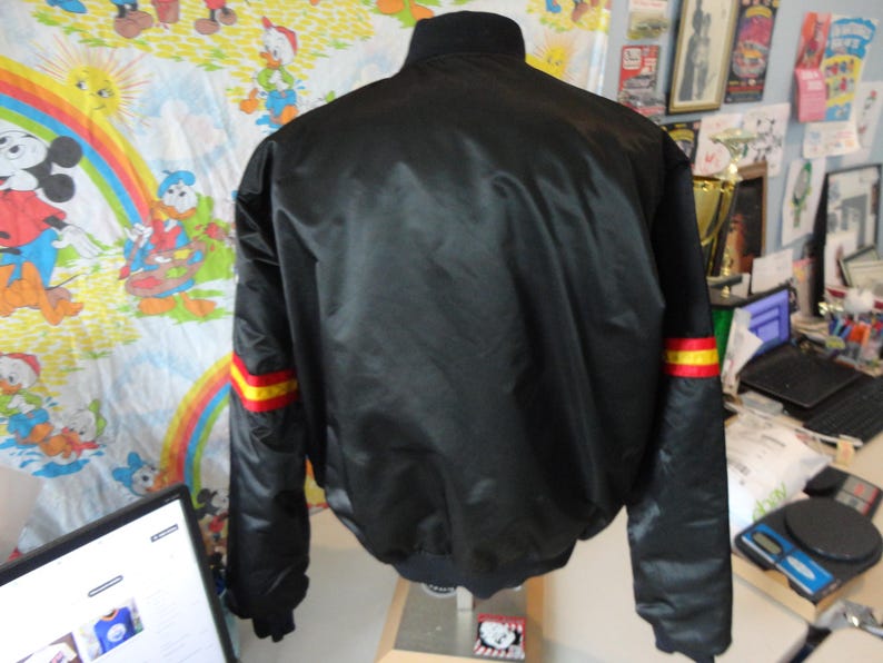 May include: Black bomber jacket with a ribbed collar and cuffs. The sleeves have red and yellow stripes near the cuffs. The jacket is made of a shiny material. The back of the jacket is visible.