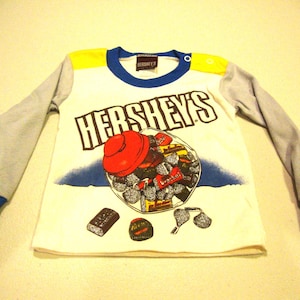 May include: A long-sleeve shirt with a white torso, light gray sleeves, and blue cuffs and collar. The front features a graphic of a glass bowl filled with Hershey's candies, the text HERSHEY'S, and individual candy illustrations including a Reese's cup.