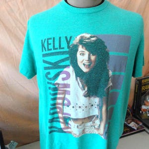 May include: Teal t-shirt featuring a graphic of a woman with dark hair and the text "Kelly Ski". The graphic is set against a purple and blue striped background. The shirt is displayed on a mannequin.