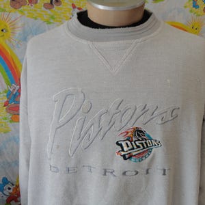 Puede incluir: Sudadera gris claro de los Pistons con el nombre y el logotipo del equipo bordados en la parte delantera. La palabra "DETROIT" está bordada debajo del logotipo. La sudadera tiene cuello redondo y mangas largas.