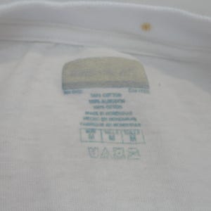 May include: Close-up of a white t-shirt collar with a label. The label includes text indicating the shirt is made of 100% cotton and made in Honduras. The label also shows washing instructions and size information.