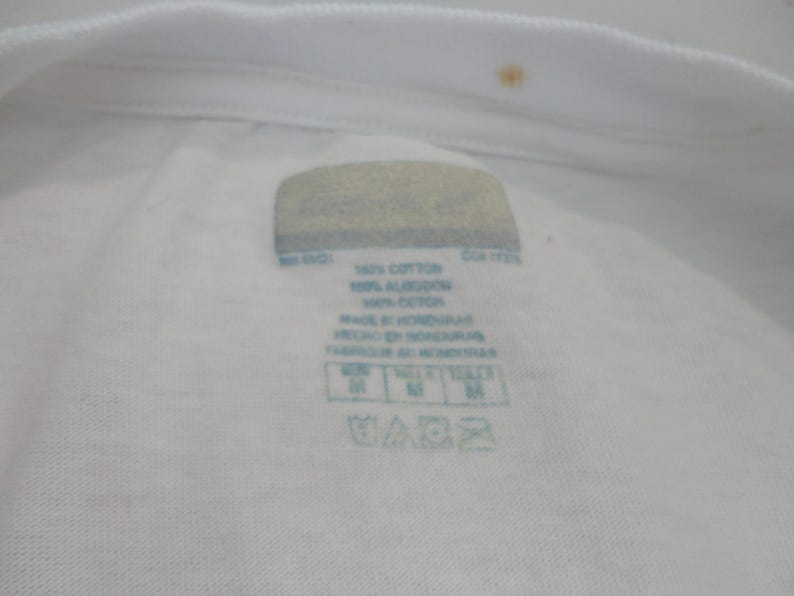 May include: Close-up of a white t-shirt collar with a label. The label includes text indicating the shirt is made of 100% cotton and made in Honduras. The label also shows washing instructions and size information.