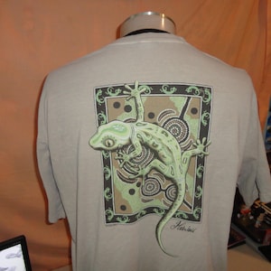 May include: A tan t-shirt featuring a detailed graphic of a green gecko with a patterned design. The gecko is inside a rectangular frame with a decorative border. The word "Hawaii" is printed below the gecko.