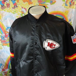 May include: Black satin jacket featuring the Kansas City Chiefs logo on the chest and NFL patch on the sleeve. The jacket has a black collar and cuffs with red and yellow stripes. A vintage sports apparel item.
