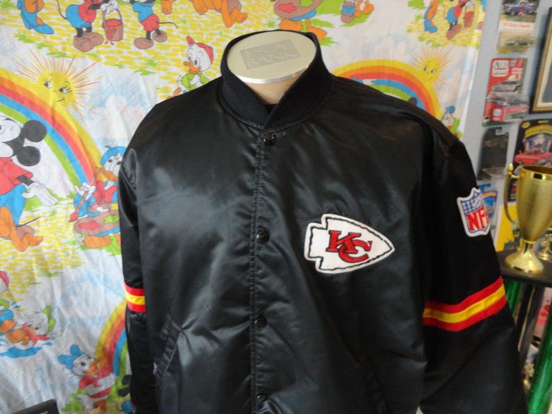 May include: Black satin jacket featuring the Kansas City Chiefs logo on the chest and NFL patch on the sleeve. The jacket has a black collar and cuffs with red and yellow stripes. A vintage sports apparel item.
