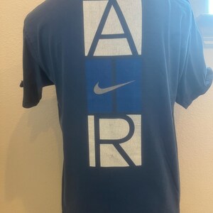 Puede incluir: Una camiseta azul marino con un gr&aacute;fico blanco de "AIR" en la espalda. La "I" de "AIR" es un cuadrado azul con un swoosh Nike blanco.