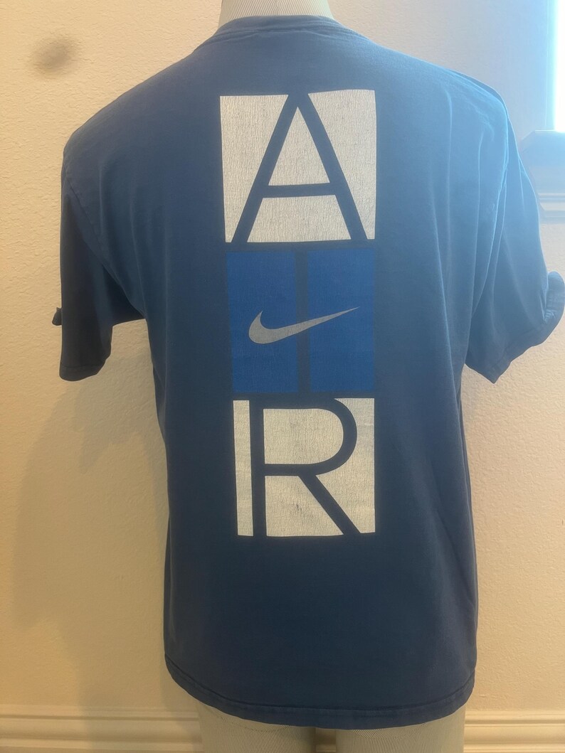 Puede incluir: Una camiseta azul marino con un gr&aacute;fico blanco de "AIR" en la espalda. La "I" de "AIR" es un cuadrado azul con un swoosh Nike blanco.