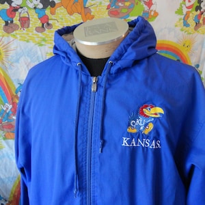 May include: A blue hooded jacket with a zipper closure and an embroidered Kansas Jayhawks logo. The logo features a blue bird with the text "KU" and "KANSAS" underneath.