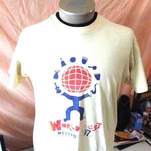 May include: Cream-coloured t-shirt with a graphic featuring a red and white globe held by a blue figure. The shirt has the text "World Fest Westport 97" in red and blue. The shirt is displayed on a mannequin.
