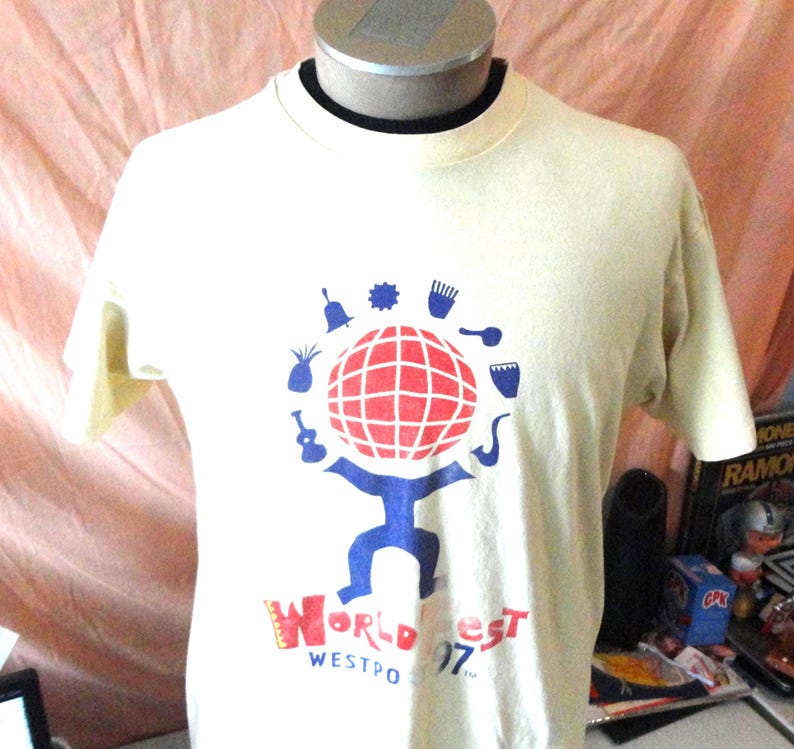 May include: Cream-coloured t-shirt with a graphic featuring a red and white globe held by a blue figure. The shirt has the text "World Fest Westport 97" in red and blue. The shirt is displayed on a mannequin.