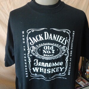 Może przedstawiać: Czarny t-shirt z białym logo Jack Daniel's Old No. 7 Brand Tennessee Whiskey. Projekt zawiera nazwę marki, rodzaj produktu i miejsce pochodzenia.