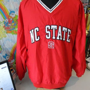 May include: Red windbreaker jacket with a white v-neck collar and white trim. The jacket has the words "NC STATE" in white letters on the front with a red and white logo.