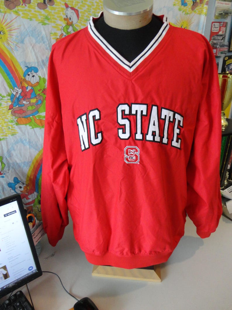 May include: Red windbreaker jacket with a white v-neck collar and white trim. The jacket has the words "NC STATE" in white letters on the front with a red and white logo.