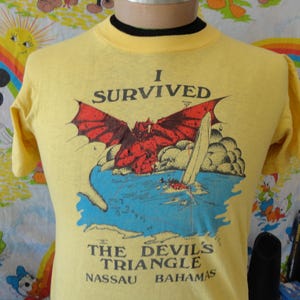 Puede incluir: Camiseta amarilla con un gráfico de un dragón rojo sobre agua azul, un velero y el texto "I SURVIVED THE DEVIL'S TRIANGLE NASSAU BAHAMAS". La camiseta tiene mangas cortas y un cuello negro.