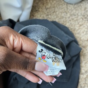 Puede incluir: Una etiqueta de camiseta negra con borde blanco. La etiqueta presenta un dibujo animado de Mickey Mouse con una camisa roja y pantalones cortos amarillos. El texto "Walt Disney World" est&aacute; impreso en blanco en la etiqueta. La etiqueta tambi&eacute;n dice "Medium" y "Hecho en Honduras".