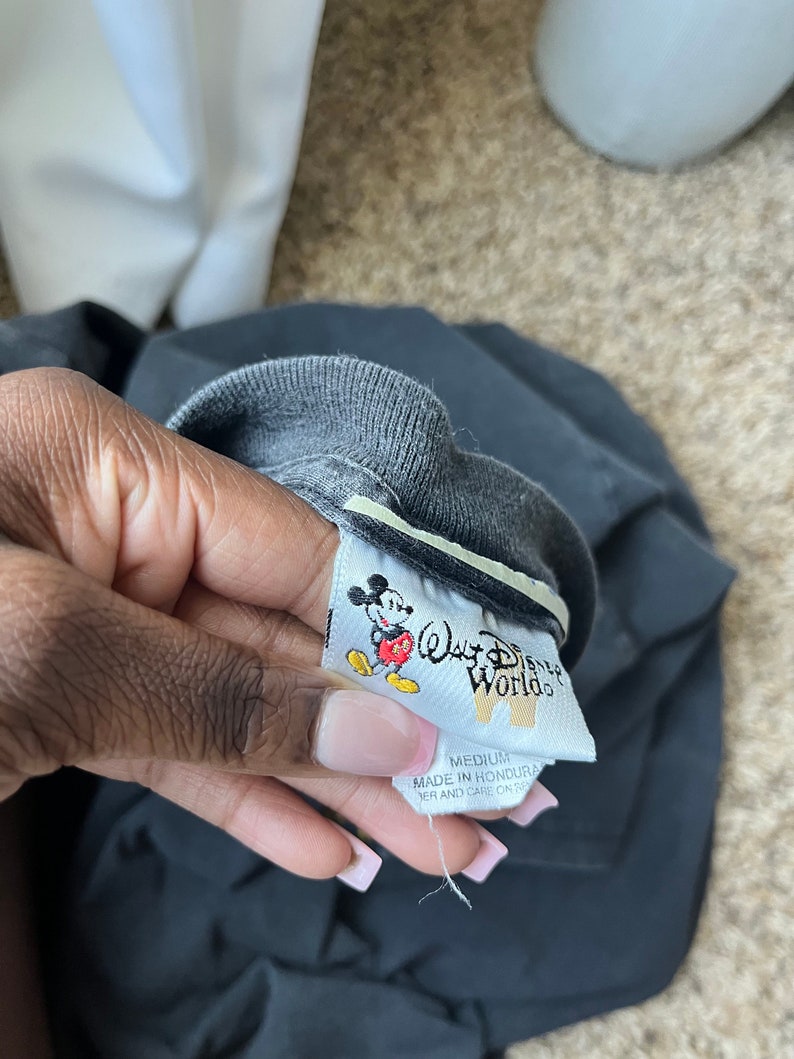 Puede incluir: Una etiqueta de camiseta negra con borde blanco. La etiqueta presenta un dibujo animado de Mickey Mouse con una camisa roja y pantalones cortos amarillos. El texto "Walt Disney World" est&aacute; impreso en blanco en la etiqueta. La etiqueta tambi&eacute;n dice "Medium" y "Hecho en Honduras".