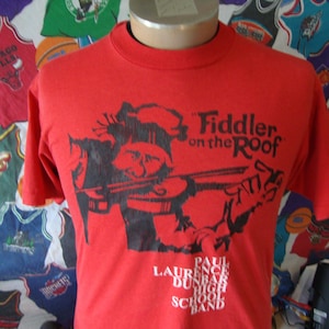 May include: Red t-shirt featuring a black and white illustration of a man playing a fiddle. The text "Fiddler on the Roof" is printed above the illustration. Below the illustration, the text "Paul Laurence Dunbar High School Band" is printed in white.