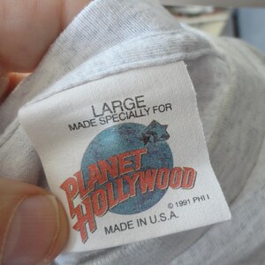 May include: A white Planet Hollywood t-shirt tag with blue and red text. The tag says "Large" and "Made Specially For Planet Hollywood". It also says "Made in U.S.A." and "&copy; 1991 PHI I".