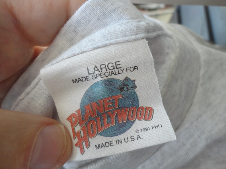 May include: A white Planet Hollywood t-shirt tag with blue and red text. The tag says "Large" and "Made Specially For Planet Hollywood". It also says "Made in U.S.A." and "&copy; 1991 PHI I".