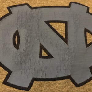 May include: A blue and black  "NC" logo on a brown background. The logo is cracked and worn.