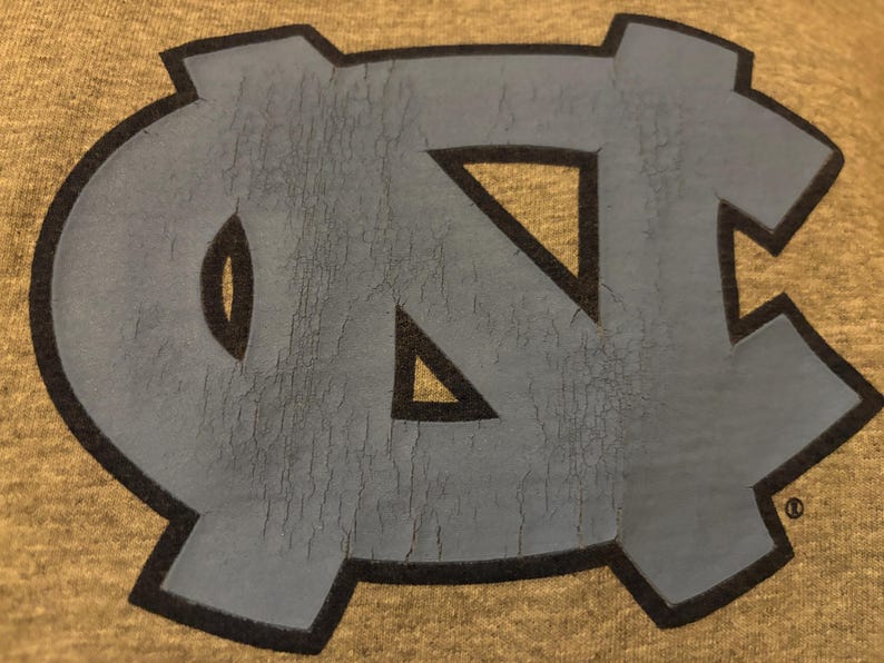 May include: A blue and black  "NC" logo on a brown background. The logo is cracked and worn.
