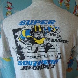 Könnte beinhalten: Ein graues T-Shirt mit einem Grafikdruck eines Footballspielers, der rennt, mit dem Text "SUPER BOWL PARTY" und "SOUTHERN REGION 7". Die Grafik enthält auch den Text "49ers vs. BRONCOS".