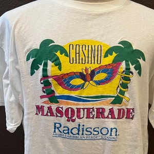 May include: White t-shirt with a colorful graphic of palm trees, a yellow sun, a masquerade mask, and the text "Casino Masquerade Radisson Aruba Caribbean Resort & Casino".