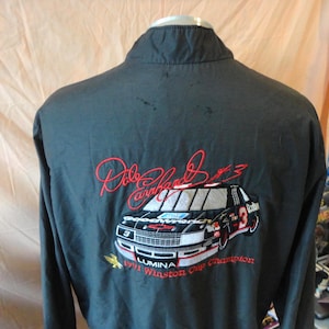 May include: Black jacket with a high collar. The back features an embroidered design of a race car with the number 3, the name "Dale Earnhardt", and the text "1991 Winston Cup Champion".