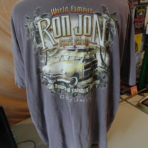 May include: A gray t-shirt featuring the "Ron Jon Surf Shop" logo with a vintage car graphic, palm trees, and the text "50 Years of Catchin' Waves Cozumel."