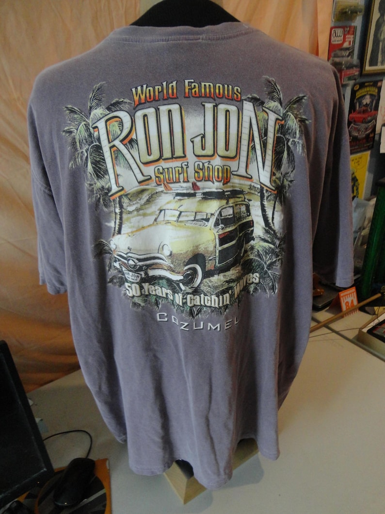 May include: A gray t-shirt featuring the "Ron Jon Surf Shop" logo with a vintage car graphic, palm trees, and the text "50 Years of Catchin' Waves Cozumel."