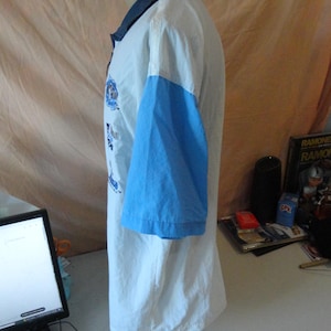 Puede incluir: Un polo de manga corta con cuerpo blanco, mangas azul claro y cuello azul oscuro. La parte delantera de la prenda muestra varios logotipos gr&aacute;ficos azules y elementos de texto dispuestos verticalmente a lo largo del pecho.
