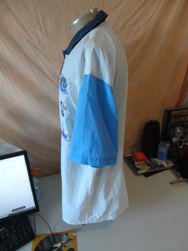 Puede incluir: Un polo de manga corta con cuerpo blanco, mangas azul claro y cuello azul oscuro. La parte delantera de la prenda muestra varios logotipos gr&aacute;ficos azules y elementos de texto dispuestos verticalmente a lo largo del pecho.