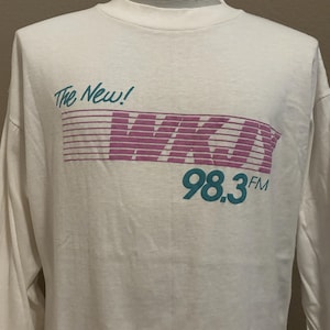 Può includere: Maglietta bianca a maniche lunghe con una grafica rosa e blu che dice "The New! WKOY 98.3 FM"