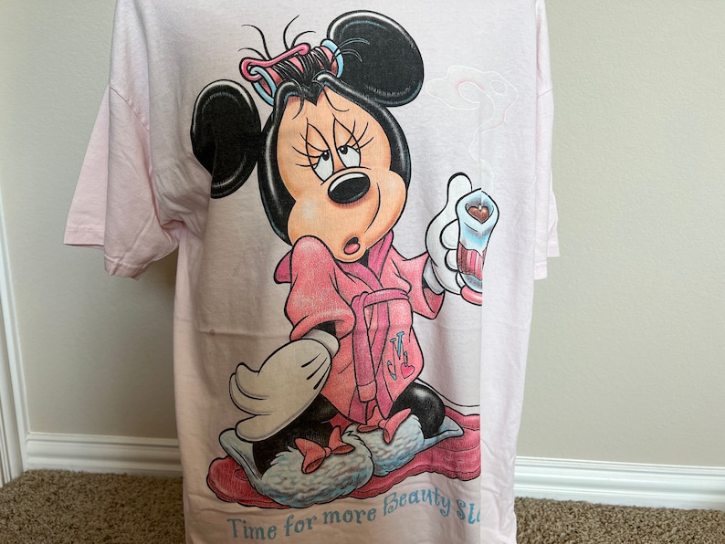 May include: Light pink t-shirt featuring a cartoon Minnie Mouse in a pink robe and curlers, holding a drink. The text "Time for more beauty sleep" is at the bottom of the shirt. The shirt is made of a soft material.