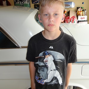 May include: A young boy wearing a black t-shirt with a baseball player graphic and the text "New York Mets".
