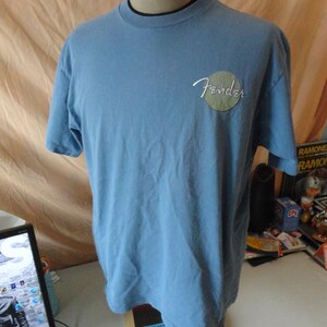 May include: Light blue Fender t-shirt with short sleeves. The Fender logo is in white script inside a green circle on the left chest. The shirt is displayed on a mannequin.
