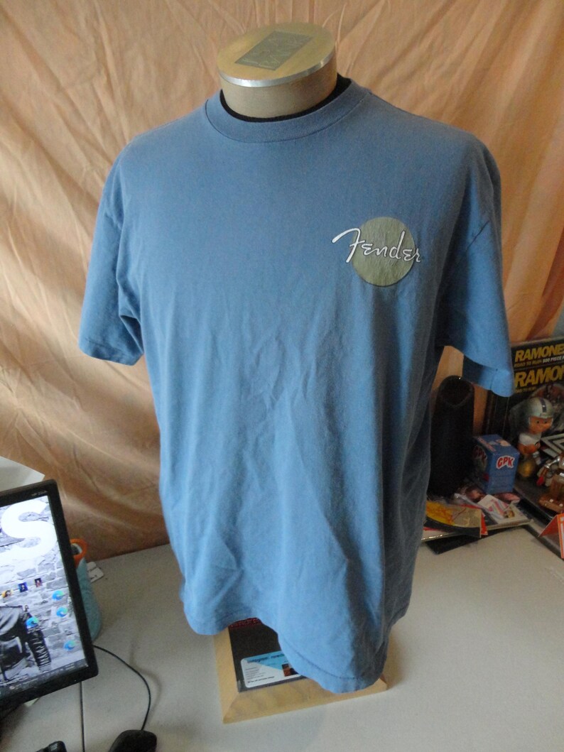 May include: Light blue Fender t-shirt with short sleeves. The Fender logo is in white script inside a green circle on the left chest. The shirt is displayed on a mannequin.