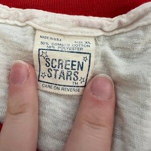 K&ouml;nnte beinhalten: Ein wei&szlig;es T-Shirt mit einem roten gerippten Kragen und einem braunen Etikett mit der Aufschrift "MADE IN USA", "SIZE XL", "50% COMBED COTTON", "50% POLYESTER", "SCREEN STARS", "TM", "CARE ON REVERSE".