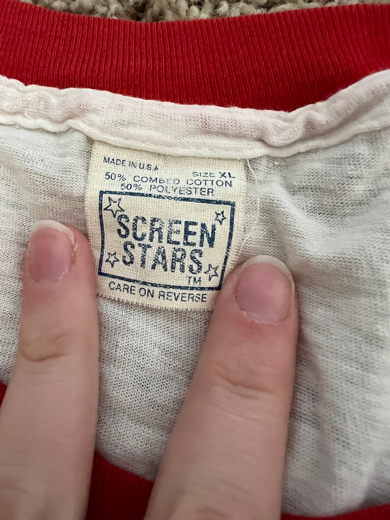 K&ouml;nnte beinhalten: Ein wei&szlig;es T-Shirt mit einem roten gerippten Kragen und einem braunen Etikett mit der Aufschrift "MADE IN USA", "SIZE XL", "50% COMBED COTTON", "50% POLYESTER", "SCREEN STARS", "TM", "CARE ON REVERSE".