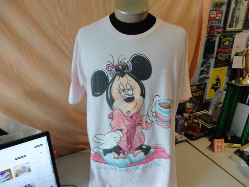 May include: A light pink t-shirt featuring a cartoon Minnie Mouse in a pink robe, holding a cup. The graphic includes the text "Time for more Beauty Sleep". The shirt is displayed on a mannequin.