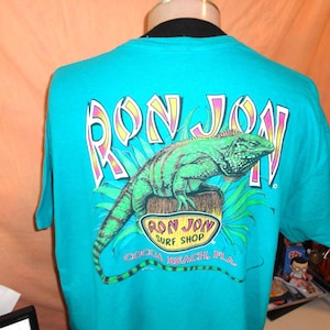 May include: Teal t-shirt with a green iguana graphic and the words "RON JON" in a colourful font. Below the iguana is the text "RON JON SURF SHOP COCOA BEACH, FLA."