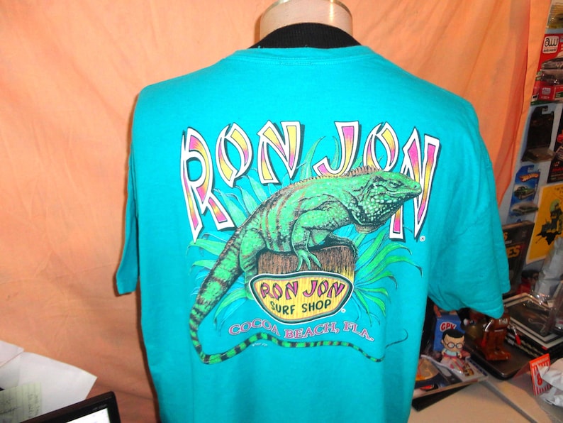 May include: Teal t-shirt with a green iguana graphic and the words "RON JON" in a colourful font. Below the iguana is the text "RON JON SURF SHOP COCOA BEACH, FLA."