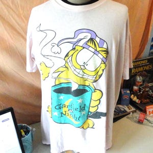 May include: A light pink short-sleeve t-shirt featuring a graphic of the cartoon character Garfield wearing a purple and white striped nightcap. Garfield is holding a teal mug with the text Garfield at Night and a yellow crescent moon.