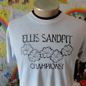 May include: Light blue t-shirt with the text "ELLIS SANDPIT CHAMPIONS!" in black, along with a floral design. The shirt has a crew neck and short sleeves. The shirt is displayed on a mannequin.